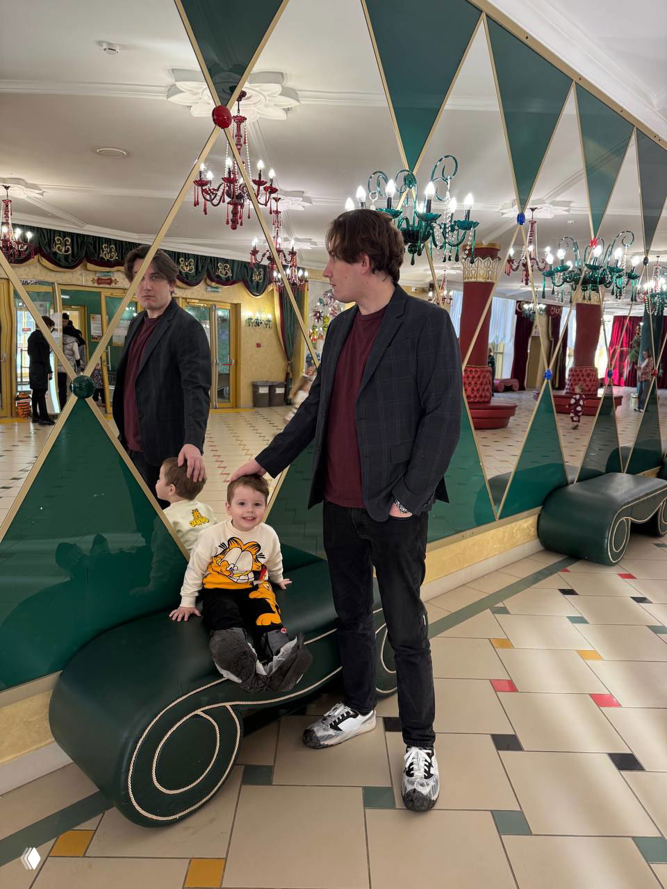 Отец рядом с ребёнком, ребёнок сидит на низкой зелёной скамье у зеркальной стены в фойе театра кукол «Экият», видна радость и доверие между ними.