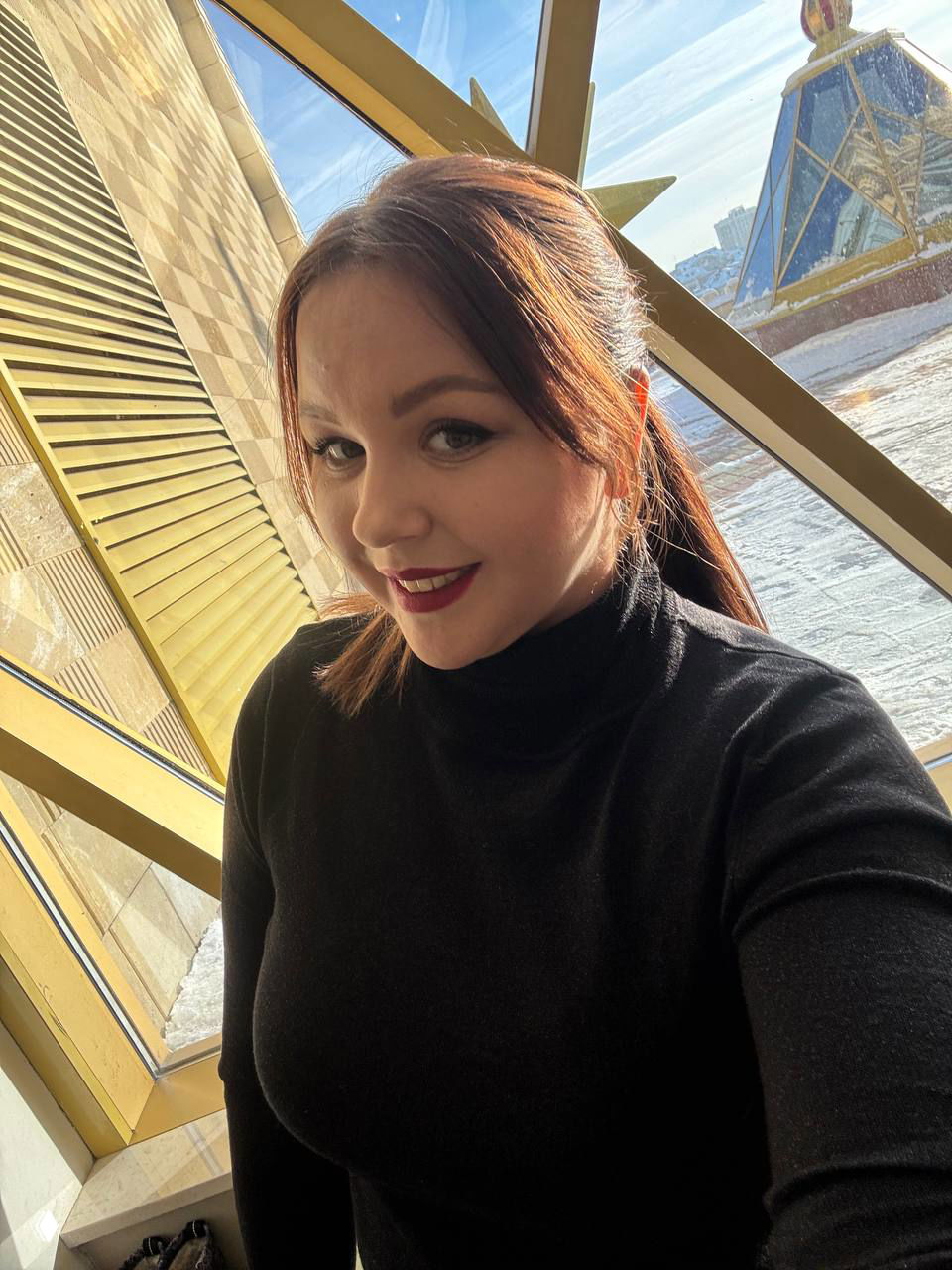 Крупный план улыбающейся мамы у окна театра, солнечный свет падает на волосы, интерьер и архитектура видны за стеклом, тёплое настроение.