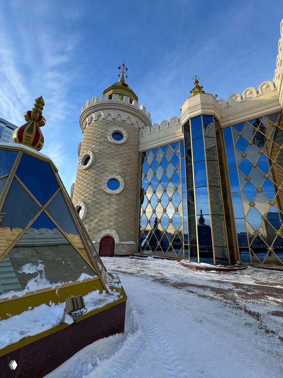 Фасад театра кукол с высокими башнями и витражными зеркальными панелями, снежная площадка перед входом и яркое голубое небо над зданием в зимний день.