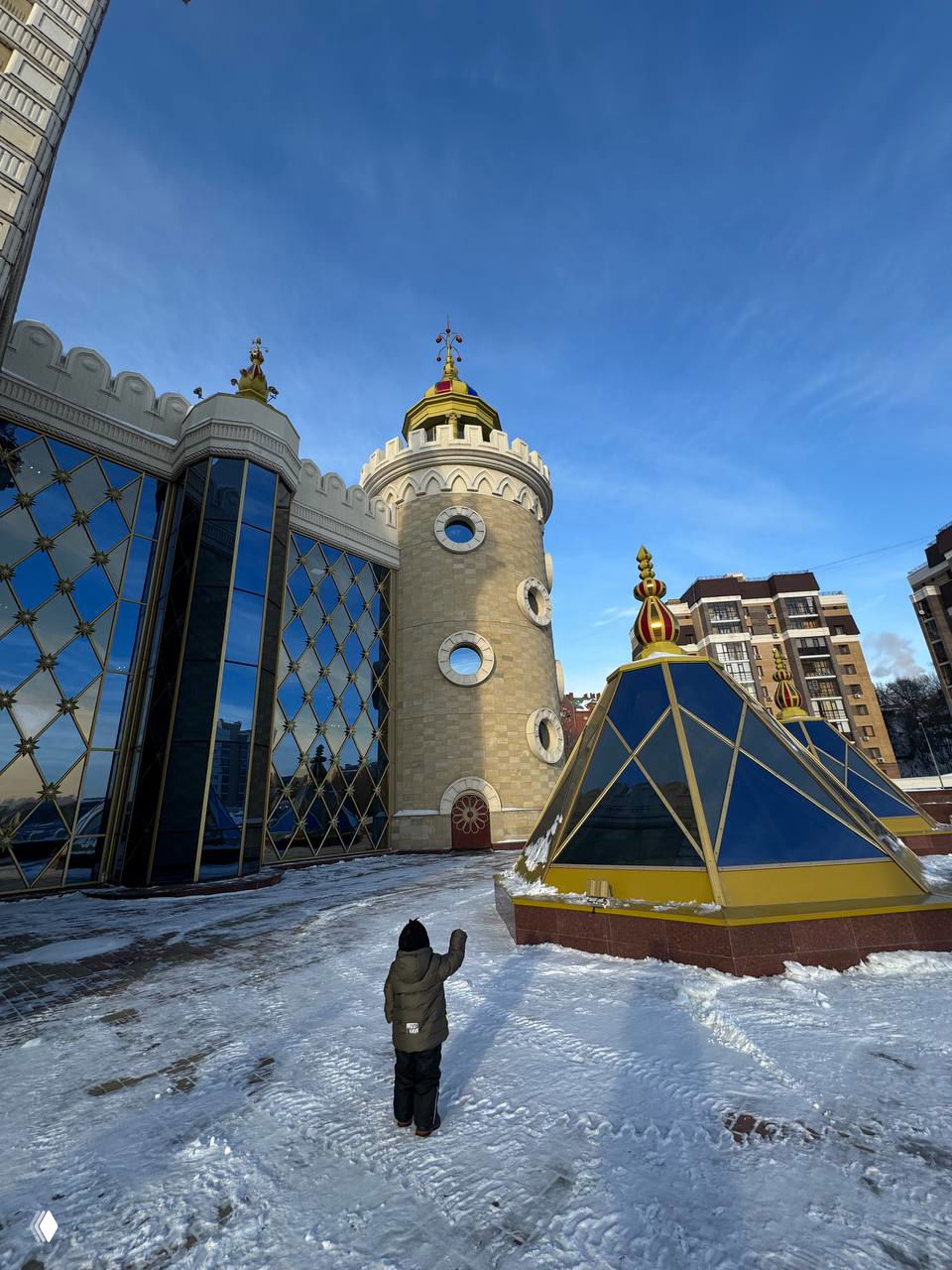 Двор и купола здания театра, сходящаяся перспектива и зимняя тропинка, ребёнок на переднем плане идёт к фасаду, подчёркивая семейный поход на представление.