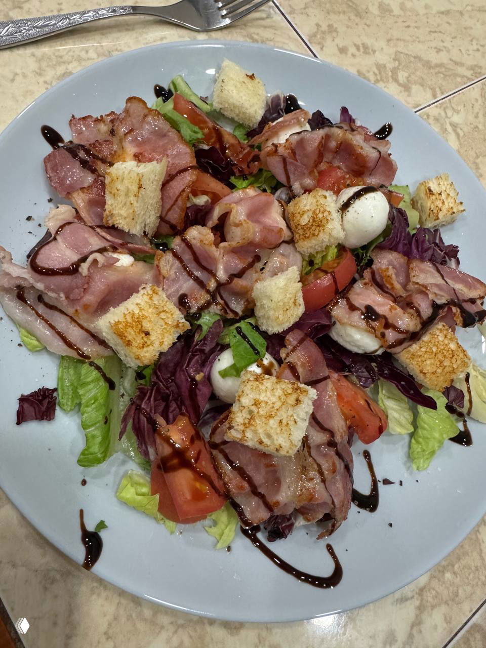 Вид сверху на тарелку с салатом: листовой микс, ломтики помидора, шарики моцареллы, несколько полосок бекона и сухарики, украшенные бальзамиком.