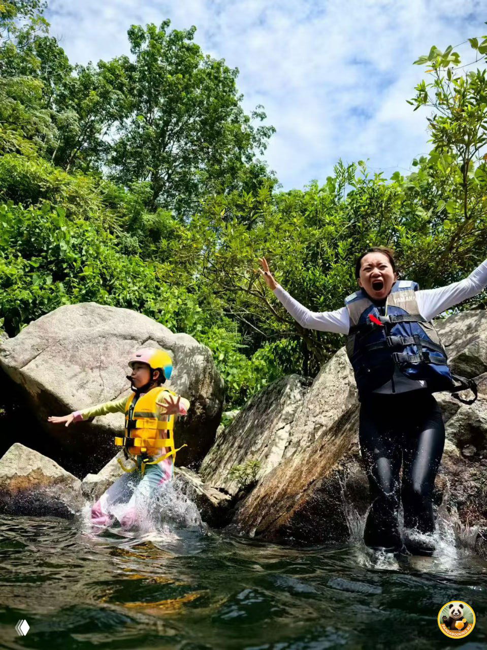 Ребёнок и взрослый в касках и спасжилетах прыгают в лесной водоём среди скал и зелёной растительности под ясным небом.