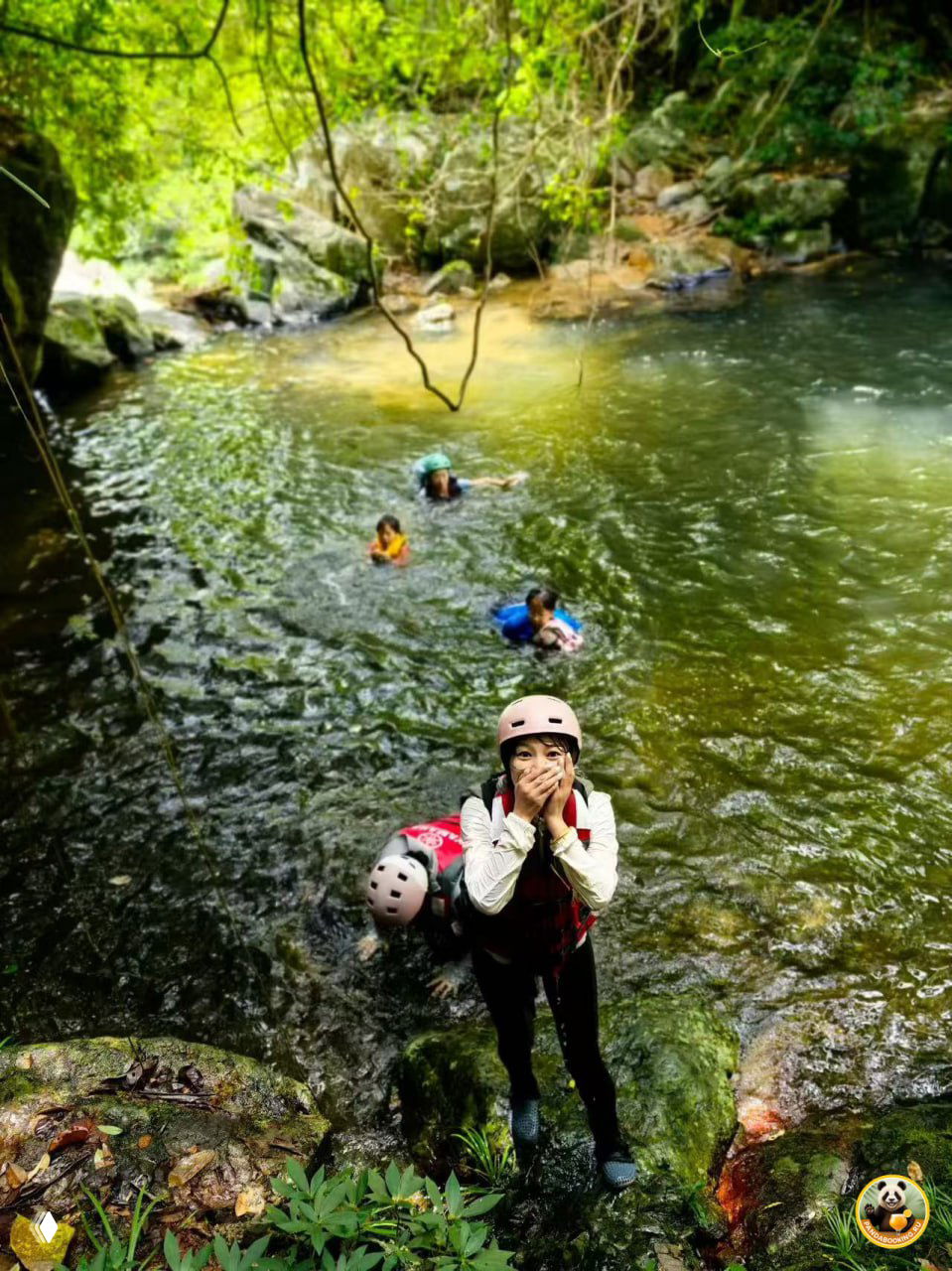 Дети в спасжилетах плывут и играют в чистом лесном бассейне; на переднем плане ребёнок стоит на скале у воды в каске.