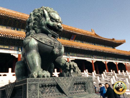 Бронзовый китайский лев-страж у входа в дворцовый комплекс, мощная скульптура на каменном постаменте на фоне традиционной кровли.