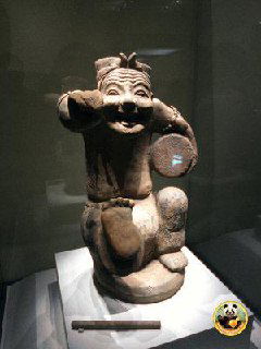 Керамическая музейная фигурка-артефакт в витрине, забавная поза и мягкое музейное освещение подчёркивают историческую ценность предмета.