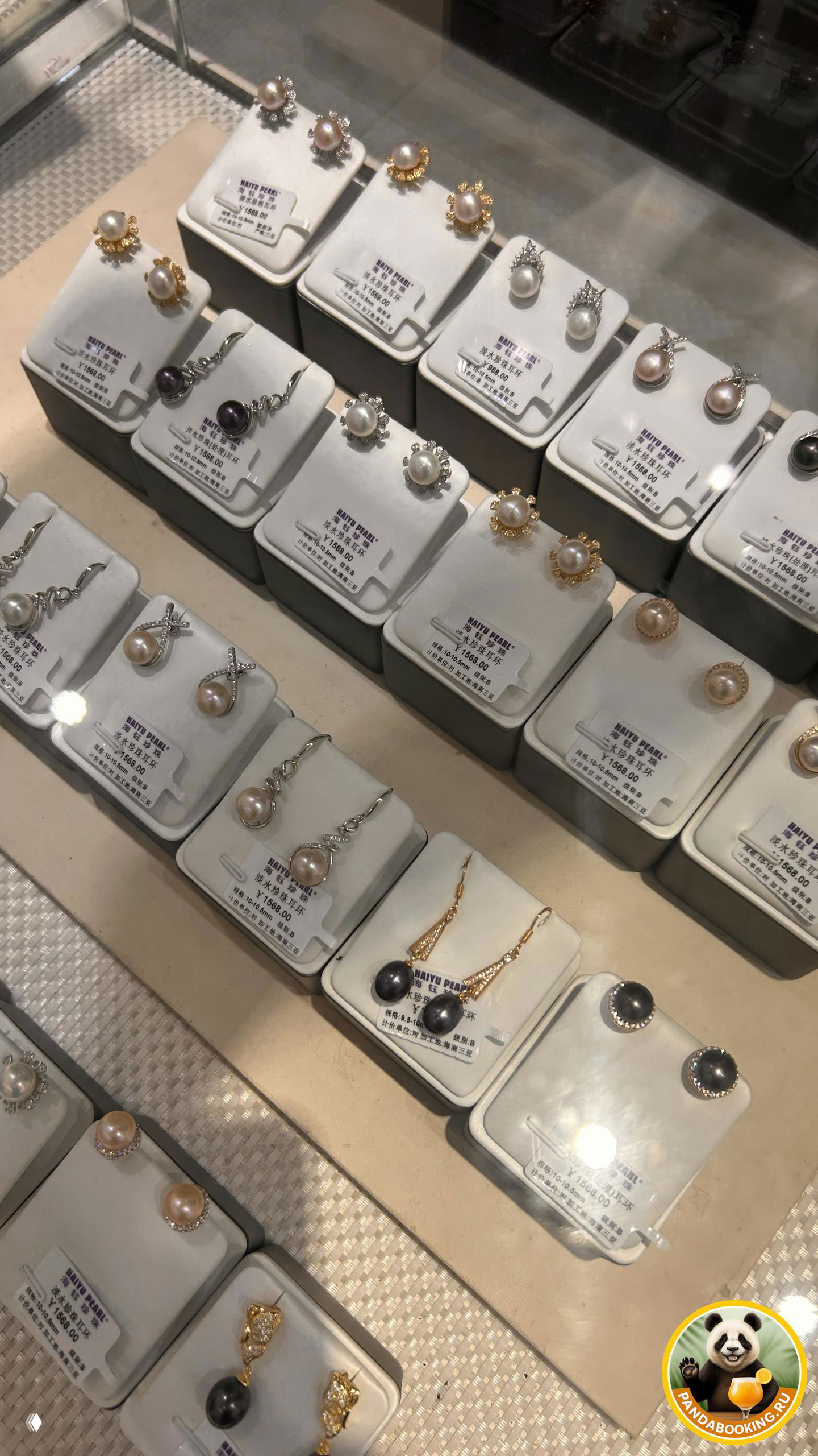 Ряд коробочек с жемчужными серьгами под стеклом, на ярлыках указаны цены и маркировка 'HAIYU PEARL', виден сеточный фон витрины.
