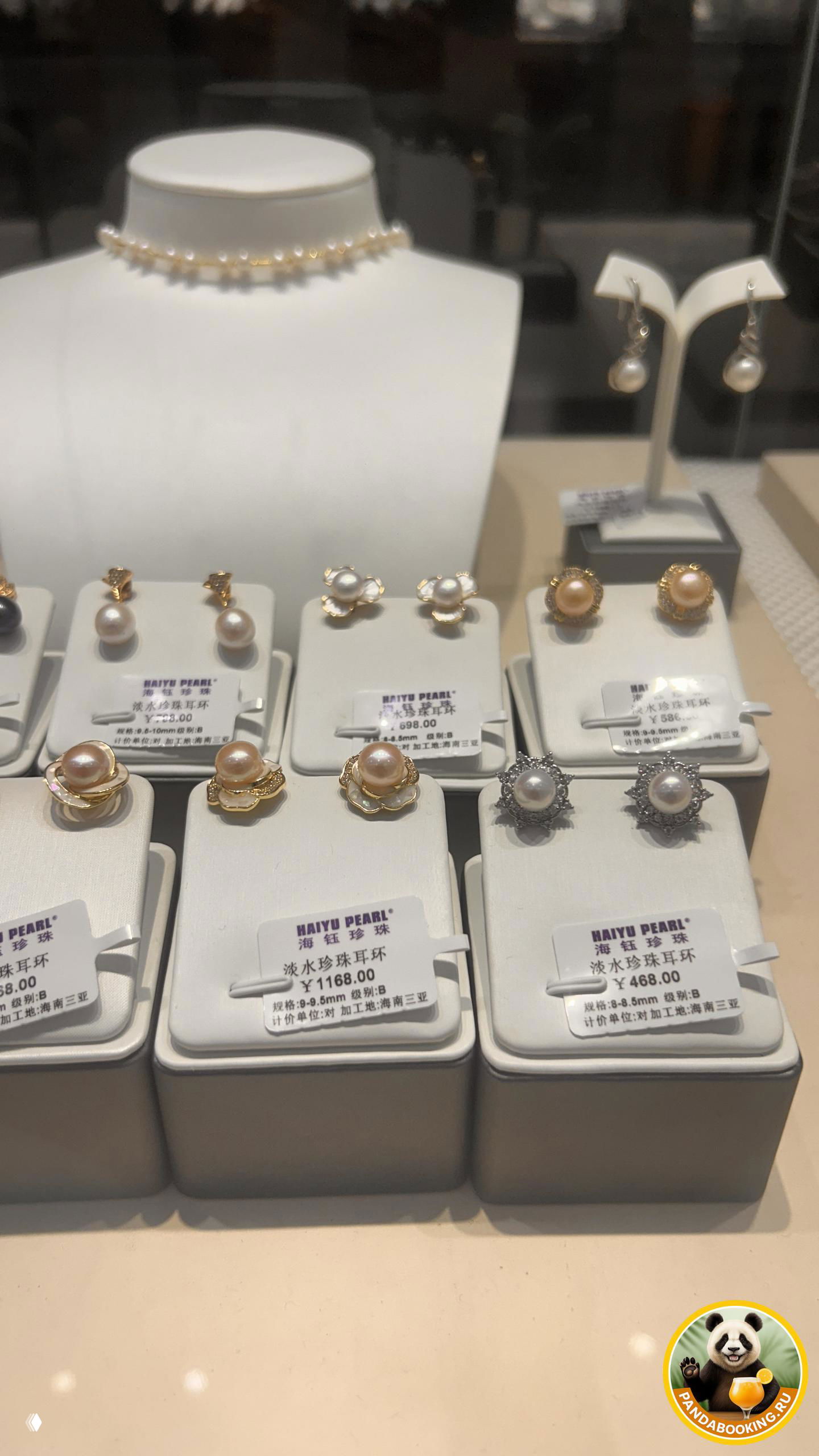 Крупный ракурс витрины с ближним планом пар жемчужных серёжек в подарочных подставках, отражение стекла и логотипа PandaBooking в углу снимка.