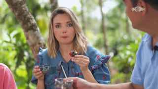 Женщина в тропическом саду пробует угощение во время съёмки телепередачи о Хайнане; видны зелёные растения, стол и микрофон на переднем плане.