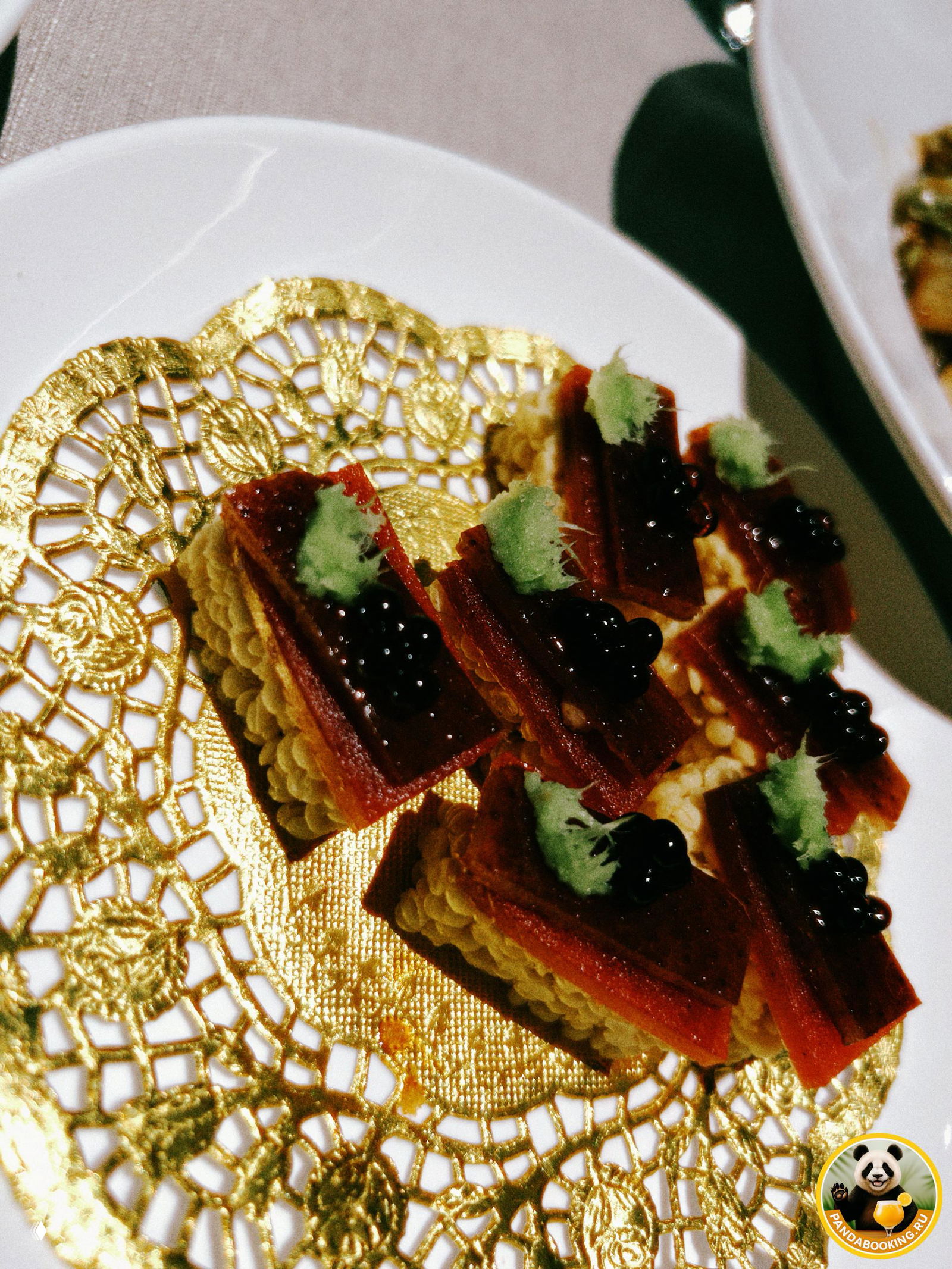 Миниатюрные закуски: слой сладкого риса, ломтик утки, соус и капля васаби на золотистой салфетке — изысканная канапе‑подача.