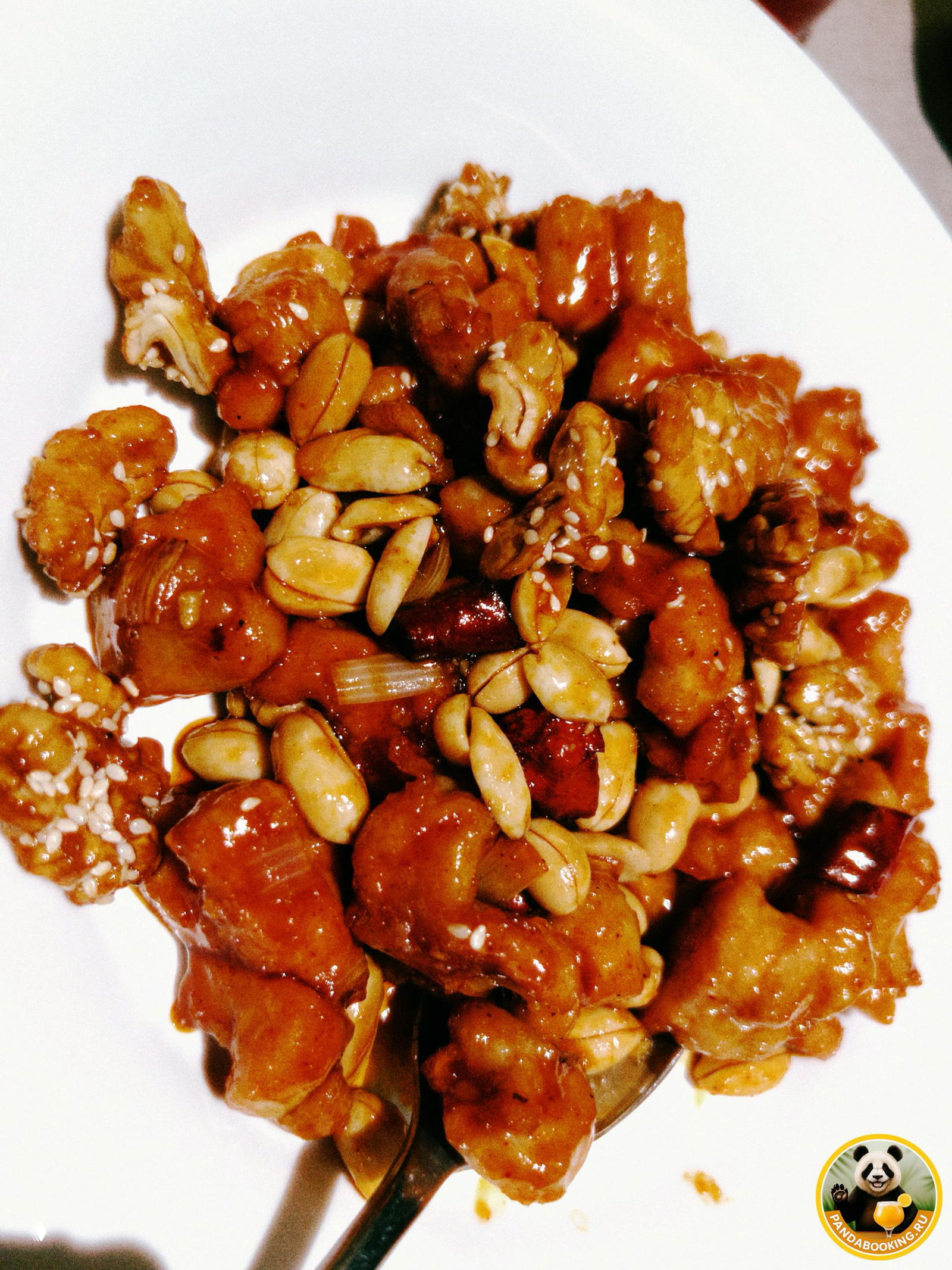 Смешение орехов и кусочков куриного мяса в карамельном соусе — классическое блюдо с арахисом и кунжутом, поданное на белой тарелке.