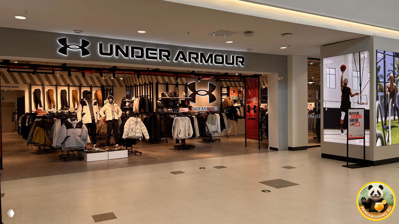 Фасад магазина Under Armour внутри Duty Free: вывеска, витрина с зимней одеждой и просторный коридор торгового уровня.