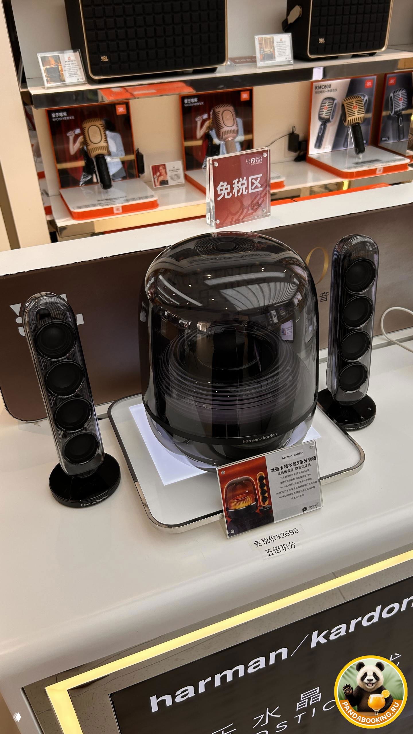 Крупный план акустической колонки Harman/Kardon на демонстрационном стенде с ценником и табличкой «免税区» — зона duty free и электроника на витрине.