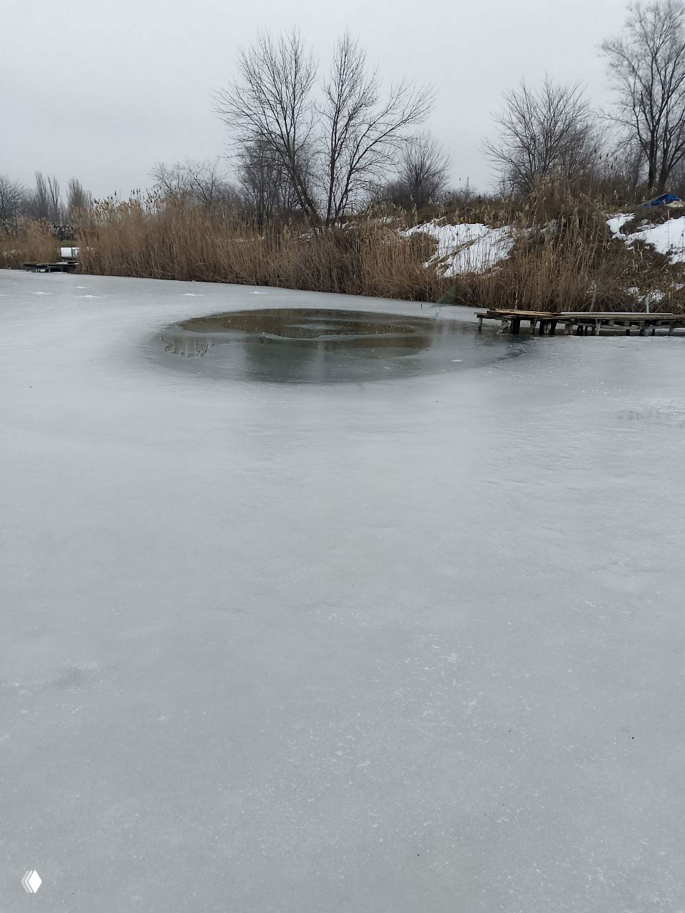 Замёрзший пруд с круглой ледовой лункой у камышовой кромки, вид с берега при пасмурной погоде, тонкий лёд и остатки снега.