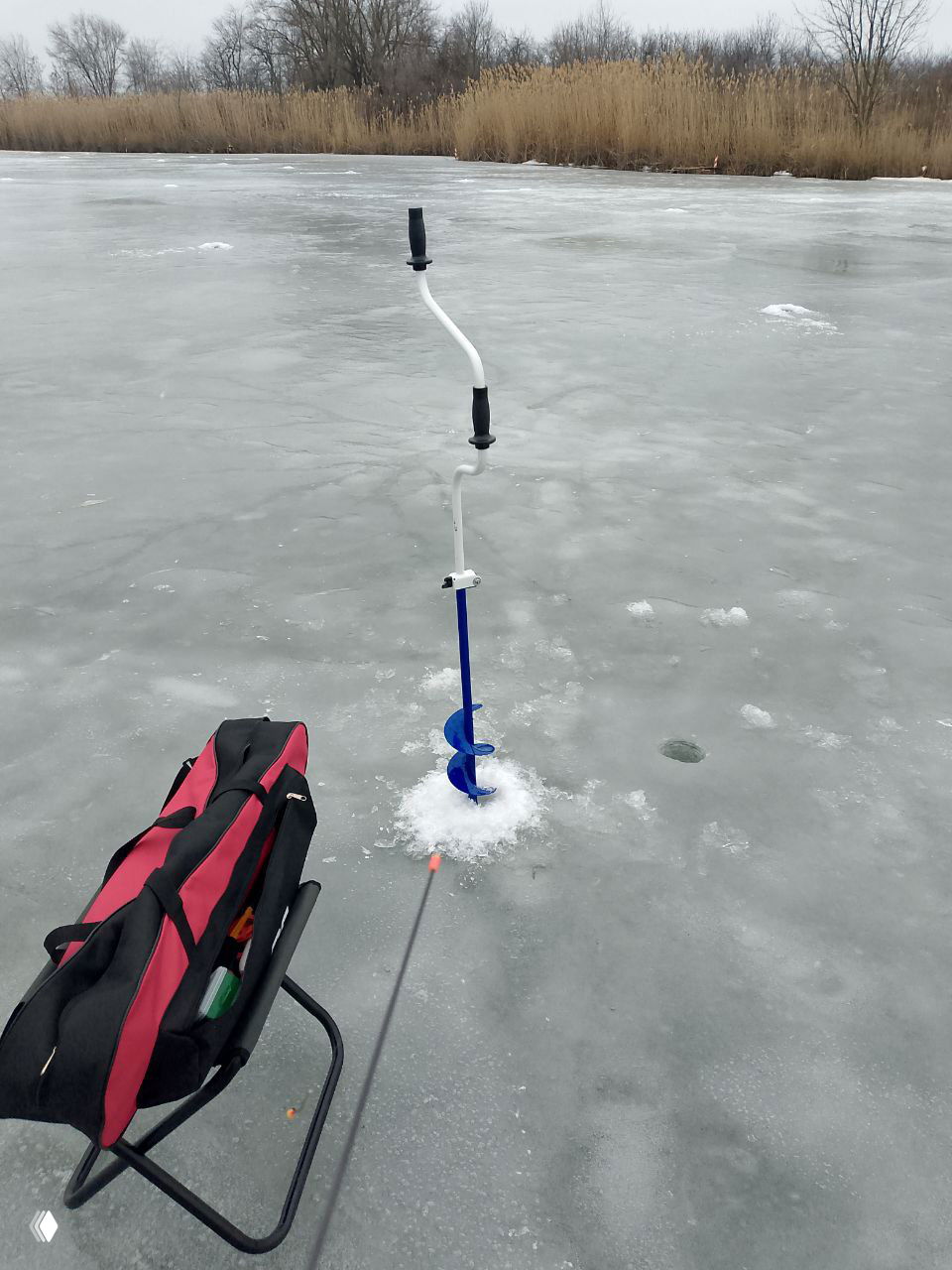 Экипировка для зимней рыбалки на льду: ярко-голубой ледобур в месте лунки, складной стул и рыболовная сумка на ледовой поверхности.