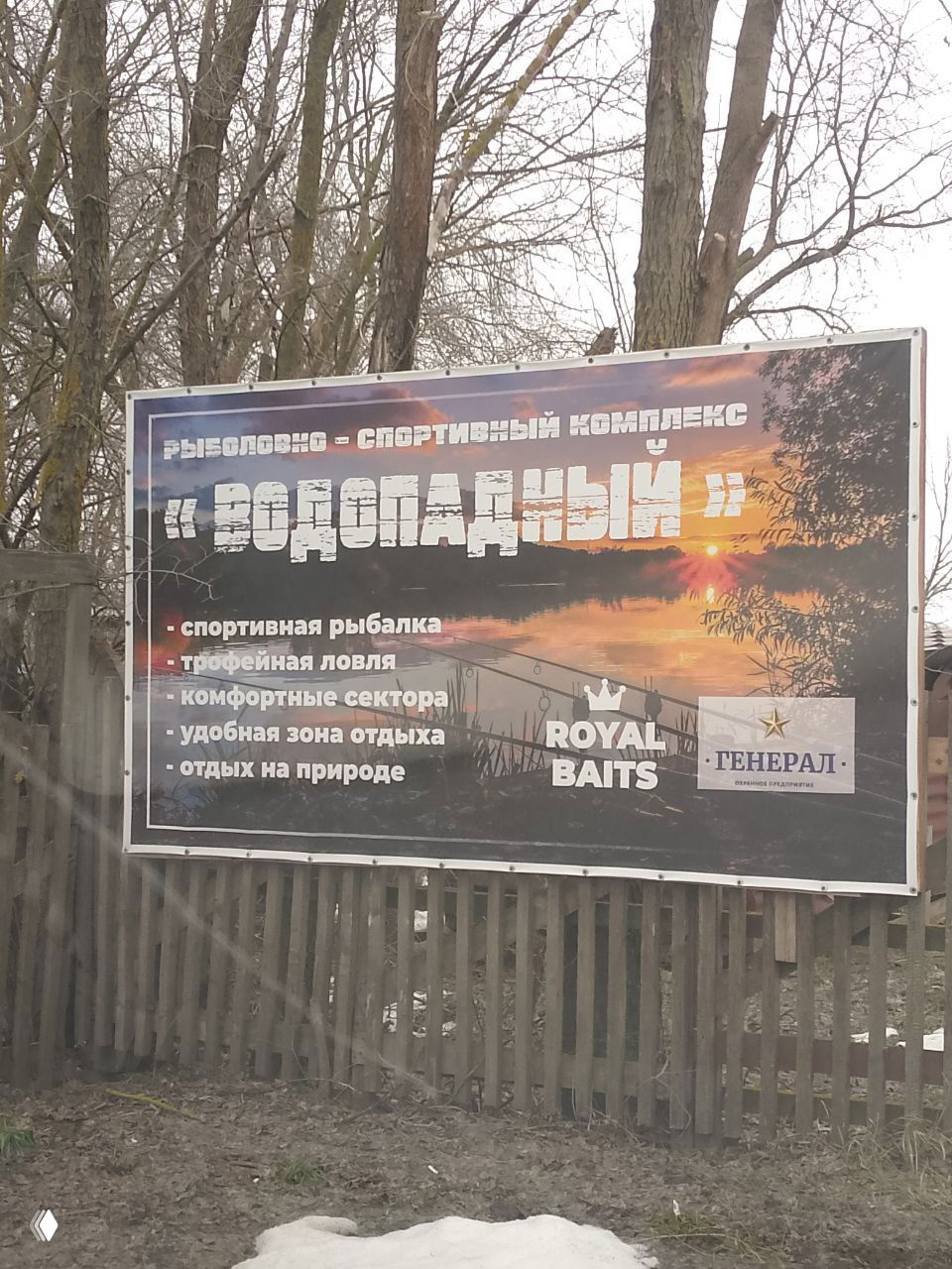 Большой информационный баннер и въездная зона рыболовно-спортивного комплекса «Водопадный» с перечнем услуг и логотипами спонсоров на фоне деревьев.