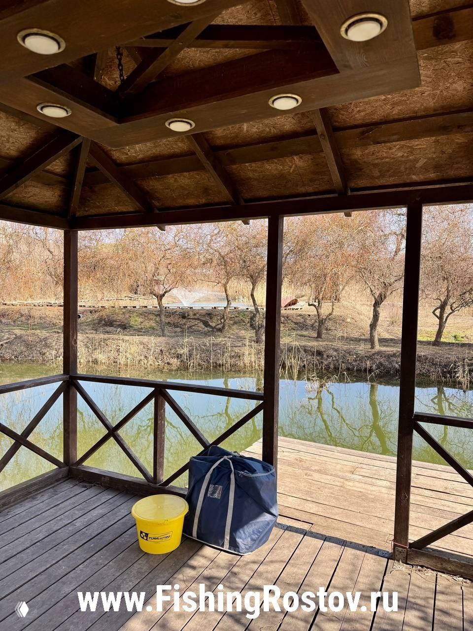 Беседка над прудом с деревянным настилом, желтое ведро и сумка для рыбалки на первом плане, спокойная поверхность воды и деревья