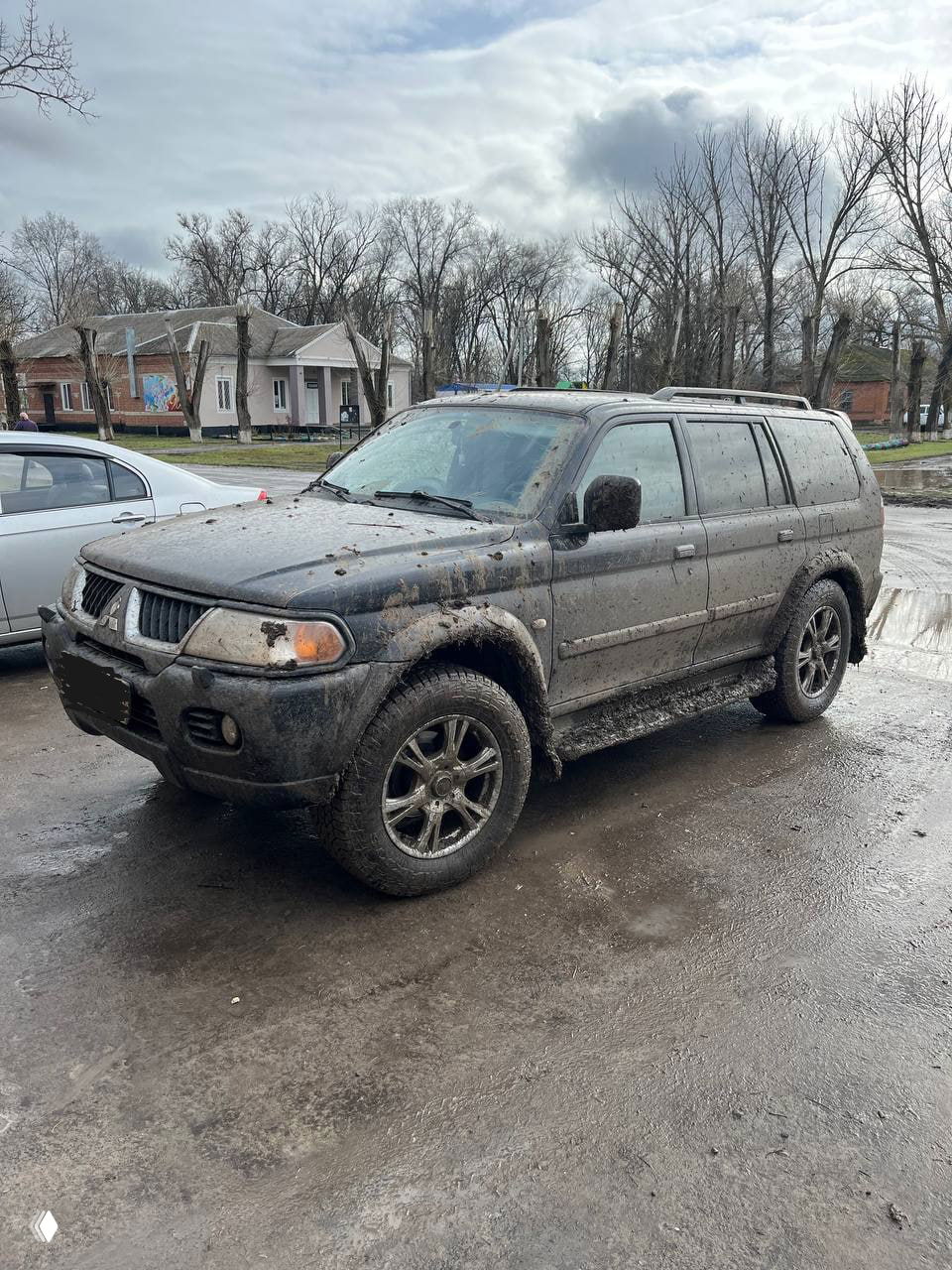 Грязный внедорожник Mitsubishi после поездки по просёлочной дороге у водоёма: брызги грязи на кузове и колёсах, деревья и дома на заднем плане.