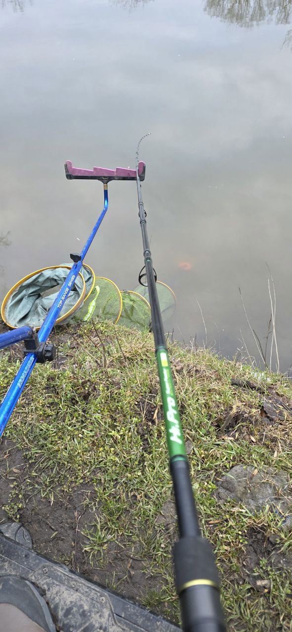 Вид на фидерное удилище, установленное на держателе, и подсак у кромки воды; тонкая режущая кромка травы у берега и отражение неба.