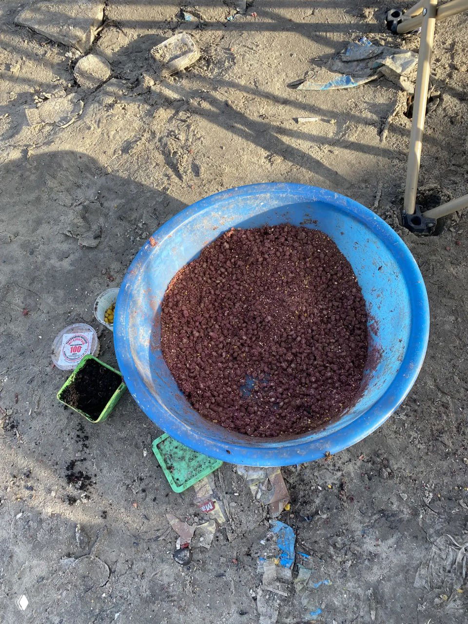 Чаша с прикормкой на песчаном берегу, рядом контейнеры с насадками и следы мусора; подготовка прикормки для донной снасти.