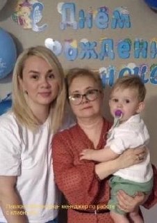 Семейная фотография с тремя людьми разного возраста и маленьким ребёнком на руках, праздничный фон с надписями и декором в помещении.