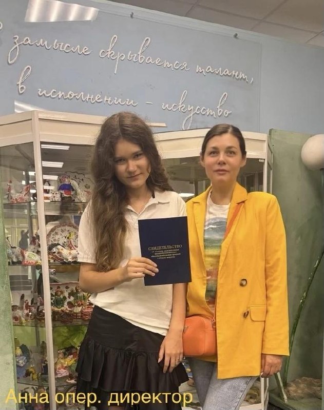 Две женщины на фоне художественной мастерской; одна держит свидетельство об окончании школы, за ними витрины с поделками и оформлением.