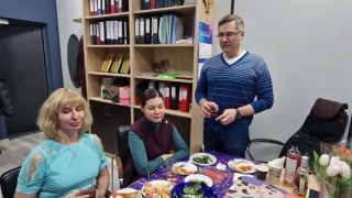 Групповое фото сотрудников у праздничного стола в офисе: три человека улыбаются, на столе блюда, напитки и весенние тюльпаны.