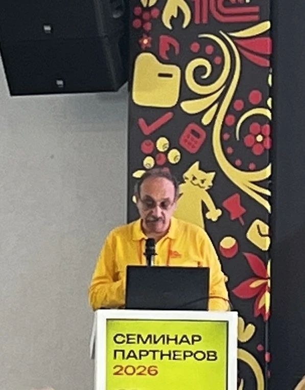 Докладчик на сцене семинара стоит за пюпитром с ноутбуком, на переднем плане табличка 'СЕМИНАР ПАРТНЕРОВ' и микрофон.