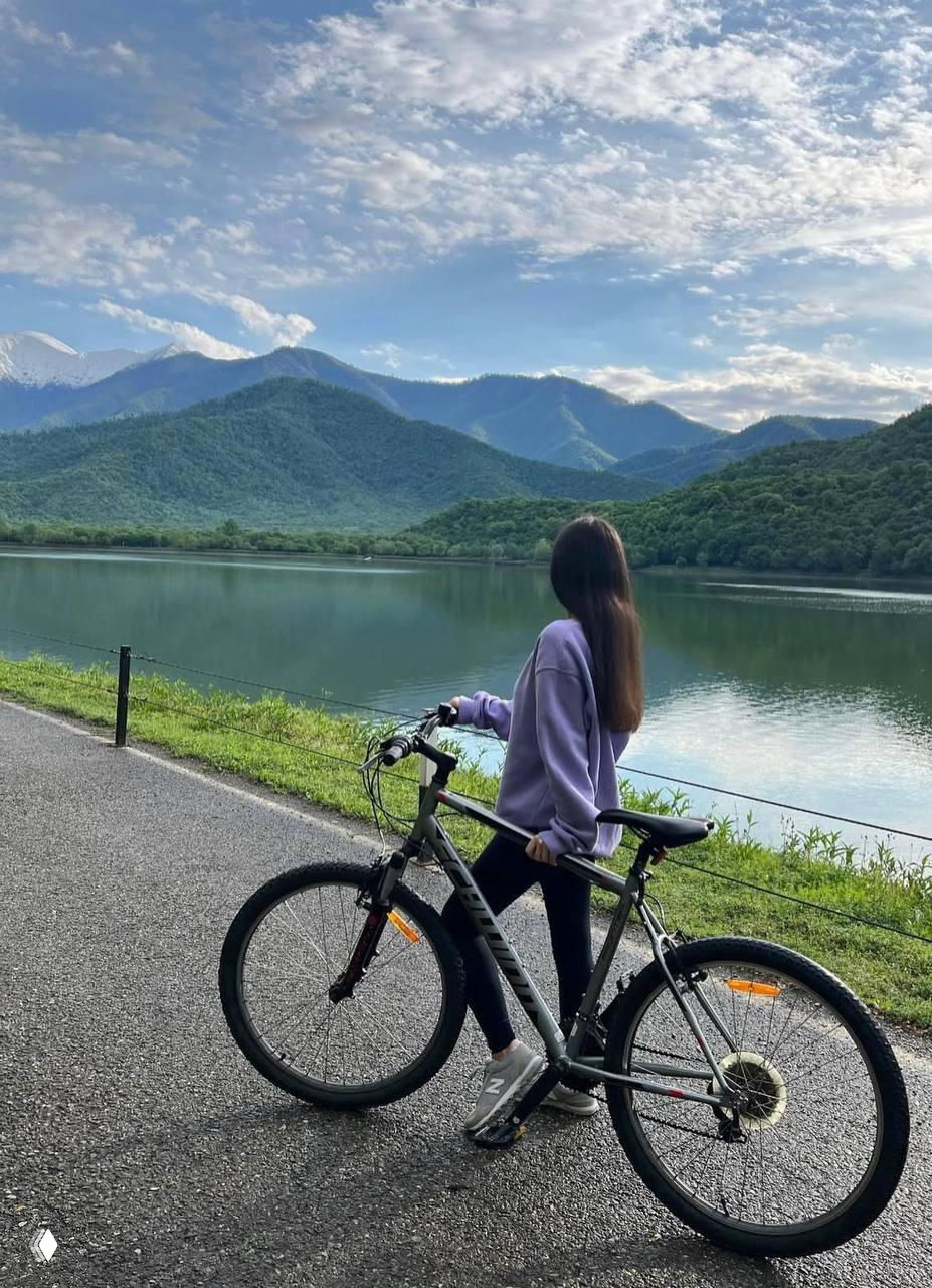 Женщина с велосипедом на асфальтовой дорожке у спокойного озера на фоне зелёных холмов и гор, отражение в воде и природный пейзаж.