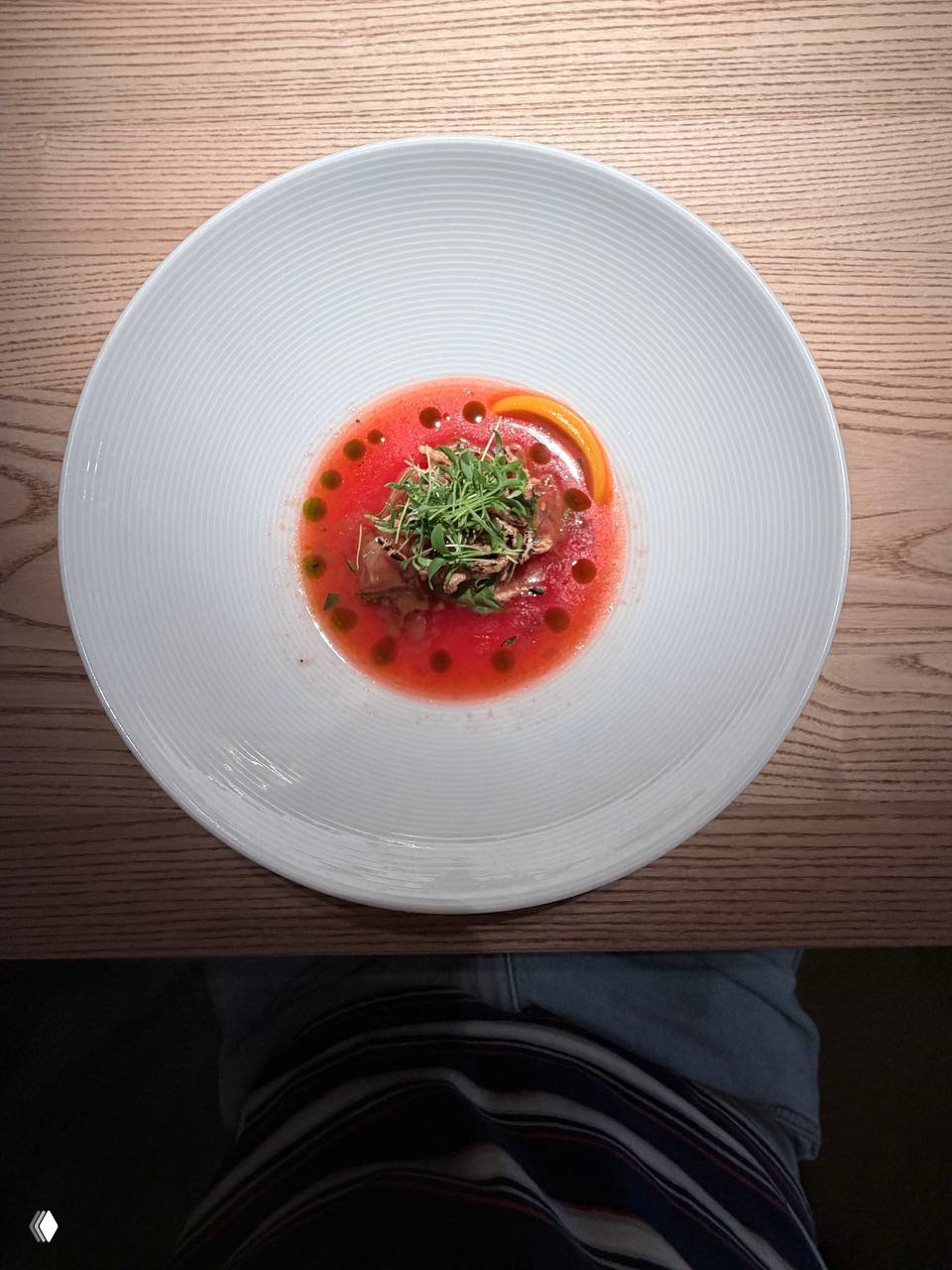 Тарелка с ярким томатным бульоном и аккуратной серединой из зелёной зелени и компонентов, подана на светлой деревянной поверхности, вид сверху.