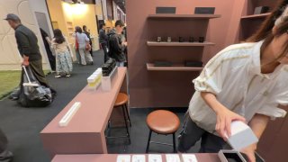 Фотография стенда с образцами парфюмов Sora Dora на выставке: полки с флаконами, стойка для тестеров и автор у столика, снимающая ролики