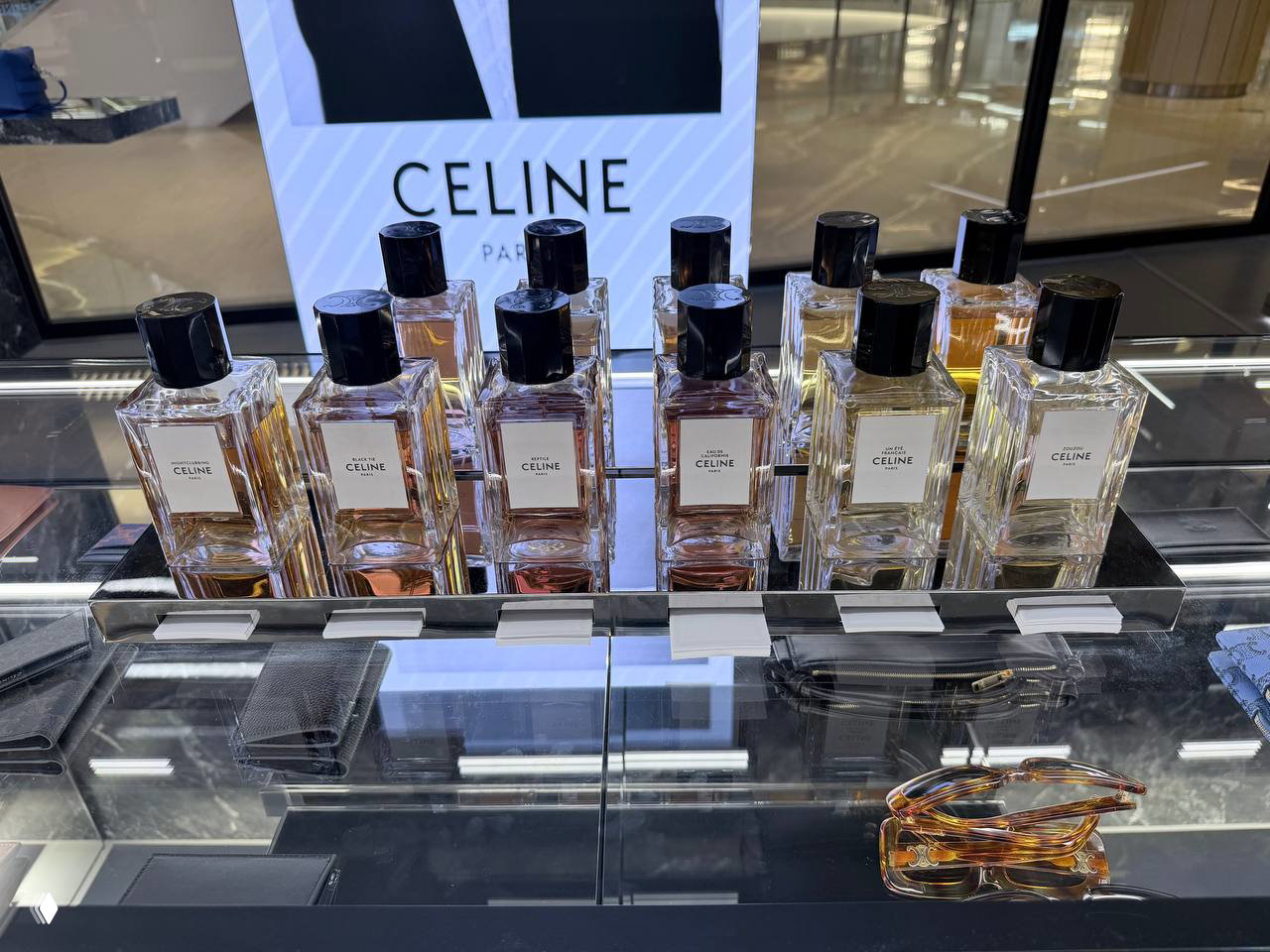 Ряд прямоугольных флаконов с белыми этикетками Celine на стеклянной стойке, отражения витрины и подсветка демонстрируют коллекцию образцов.