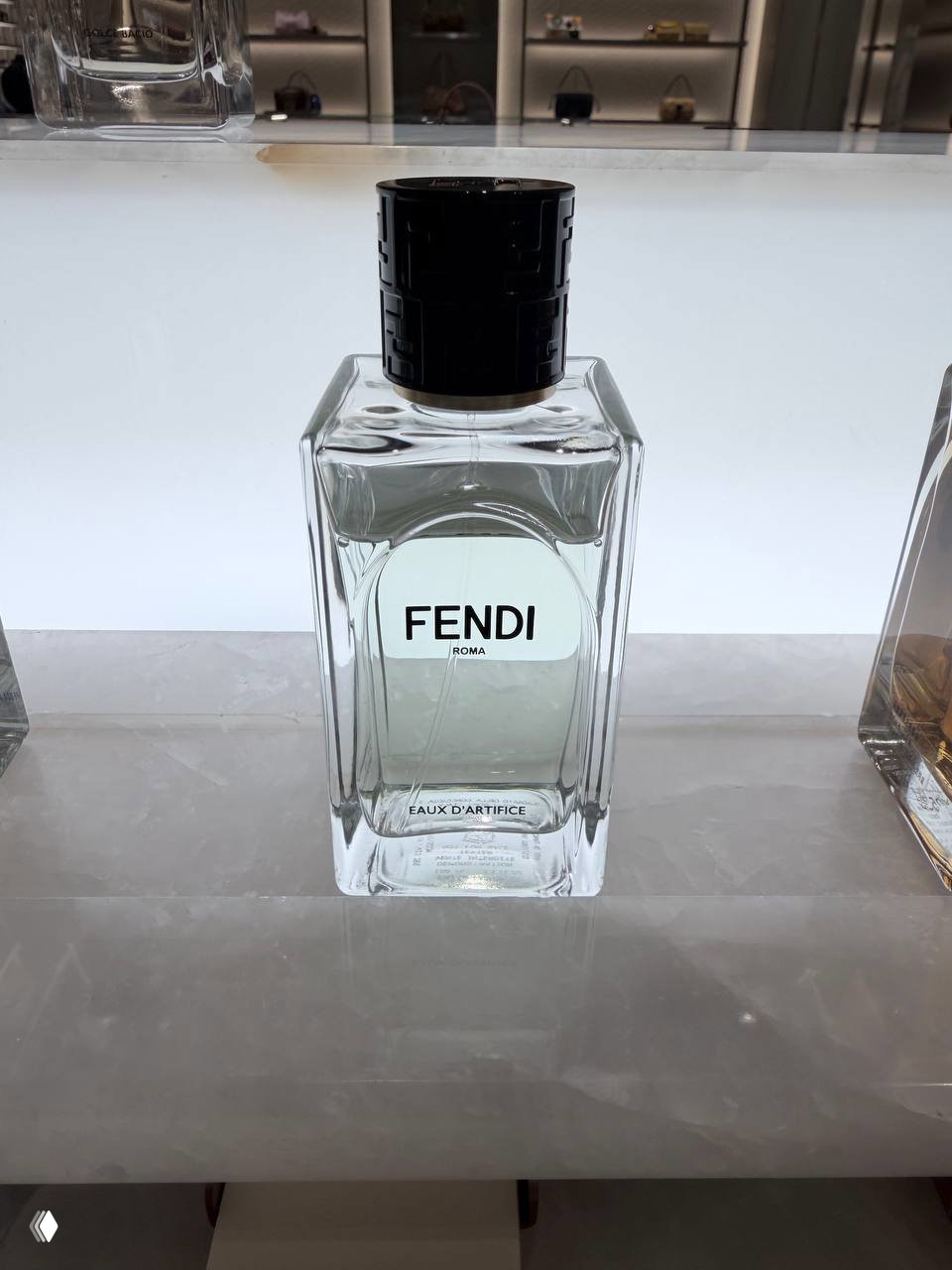 Крупный план флакона FENDI Eaux d'Artifice на светлой мраморной поверхности витрины: прозрачный стеклянный флакон с черной крышкой и логотипом.