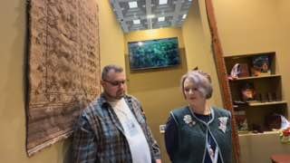 Креативный директор и собеседник в выставочном стенде на фоне ковра, витрин и жёлтой стены; кадр передаёт атмосферу презентации парфюмерной линейки.