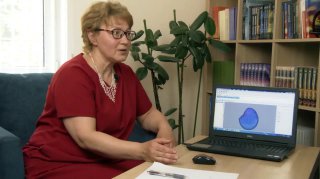 Женщина в кабинете рядом с ноутбуком, на экране видна цветная визуализация аурального электромагнитного поля; на фоне книги и растение.