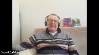 Мужчина в наушниках во время онлайн-интервью на фоне полки с книгами; кадр в домашней, спокойной обстановке, виден торс и улыбка