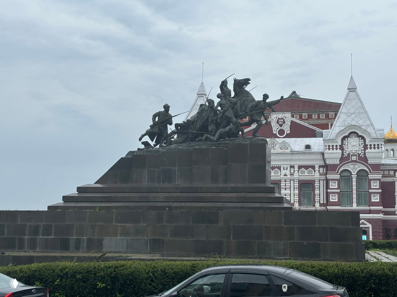 Большой бронзовый монумент с группой фигур на высоком пьедестале, на фоне исторического здания и пасмурного неба в центре Самары.