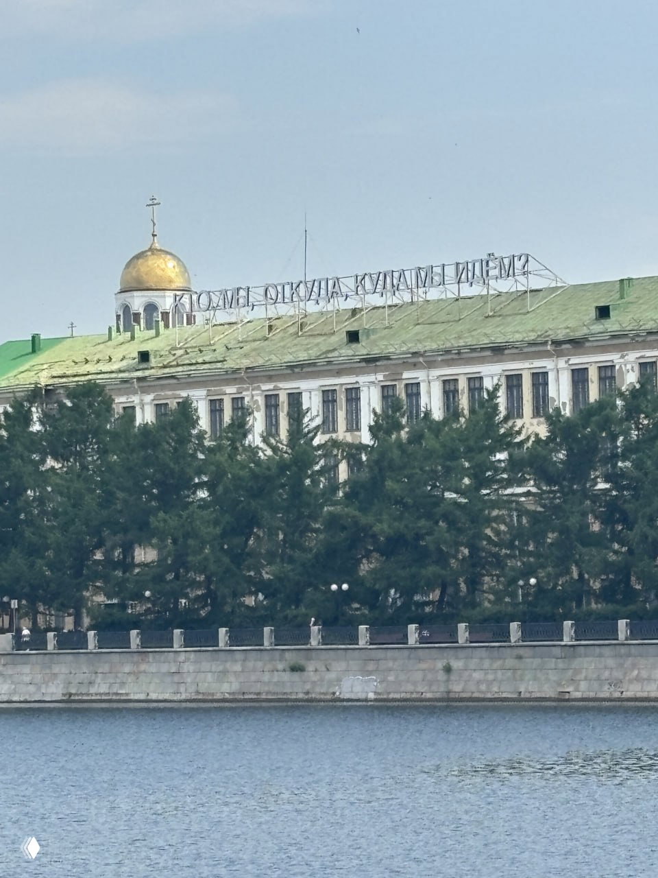 Вид с набережной Исети на береговую линию: дом с золотым куполом и большой металлической вывеской на крыше, деревья вдоль воды.