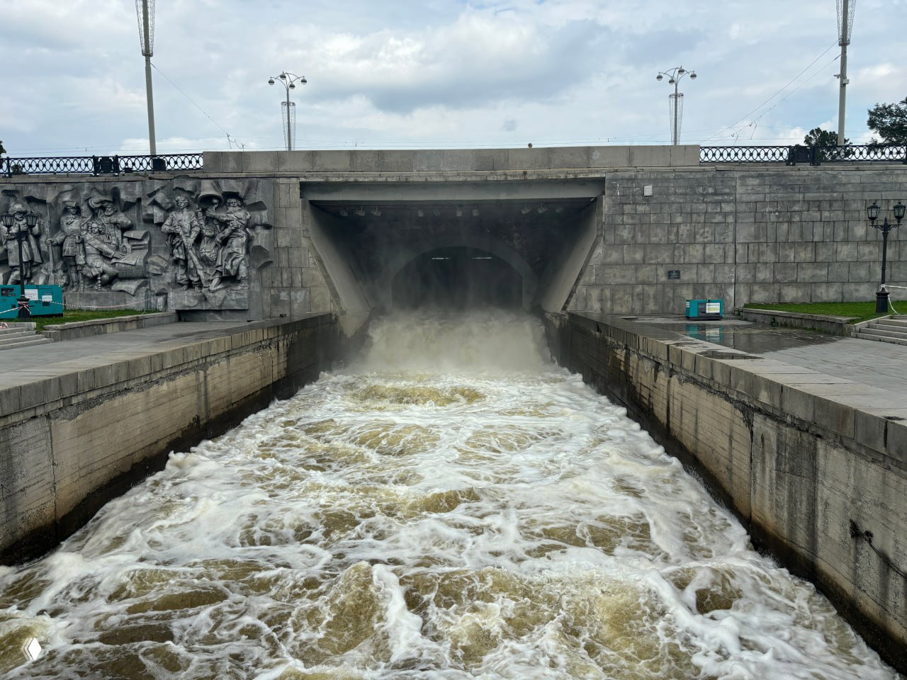 Бурный поток воды через шлюз на Плотинке: стремительная струя между бетонными стенами, над тоннелем видна скульптурная рельефная композиция.