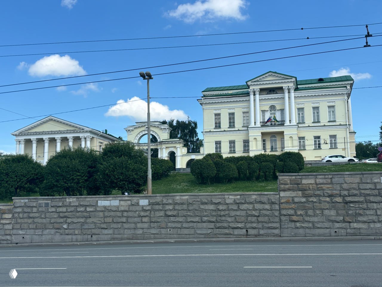 Исторические фасады с колоннами вдоль набережной Екатеринбурга: пастельные здания, стриженные кусты и дорожка у реки на переднем плане.