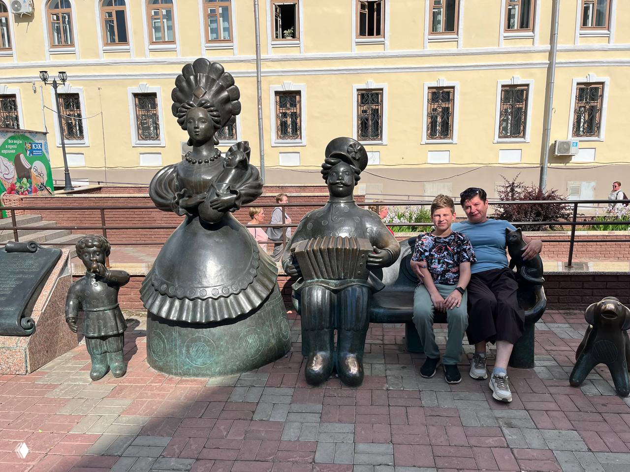 Группа бронзовых фигур и скамья в центре Кирова, рядом люди позируют для фотографий в солнечный день на городской площади.