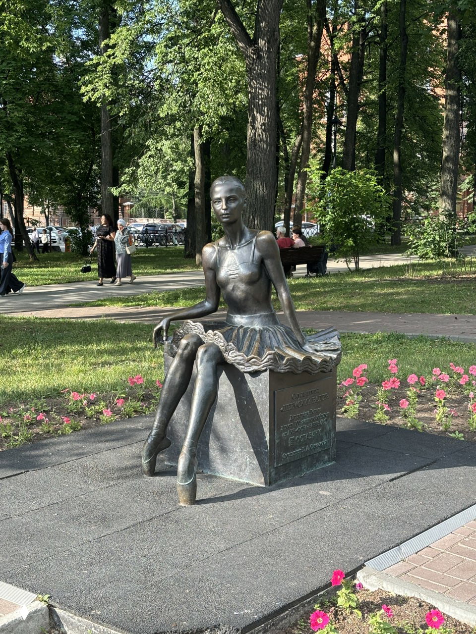Скульптура балерины в парке на фоне деревьев и дорожек, памятник в натуральную величину окружён зеленью и цветами летним днём в Перми