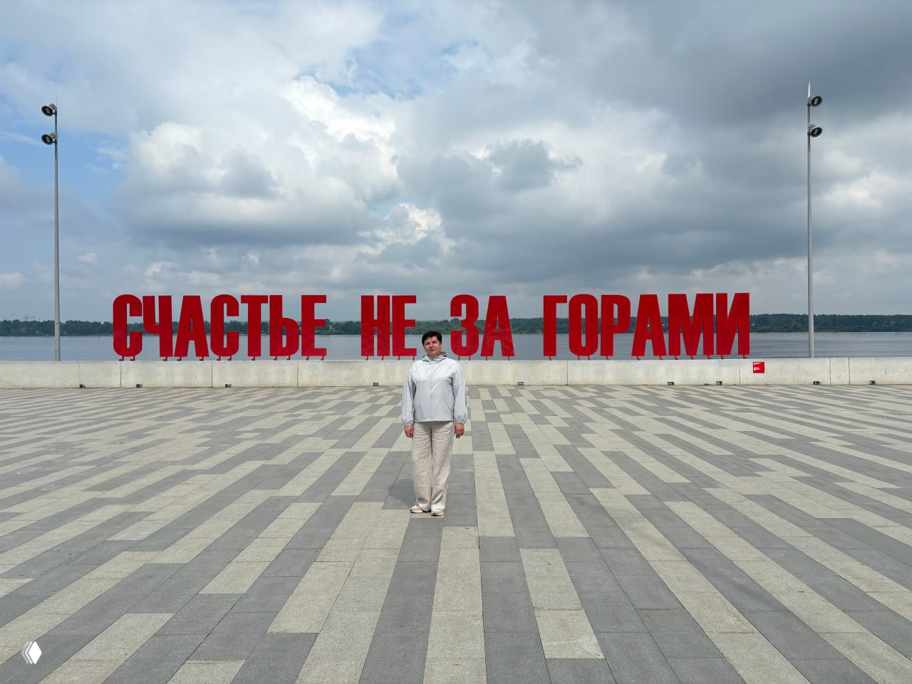 Крупная инсталляция «Счастье не за горами» на набережной Камы, большой красный текст на фоне реки и облачного неба, пешеход на переднем плане