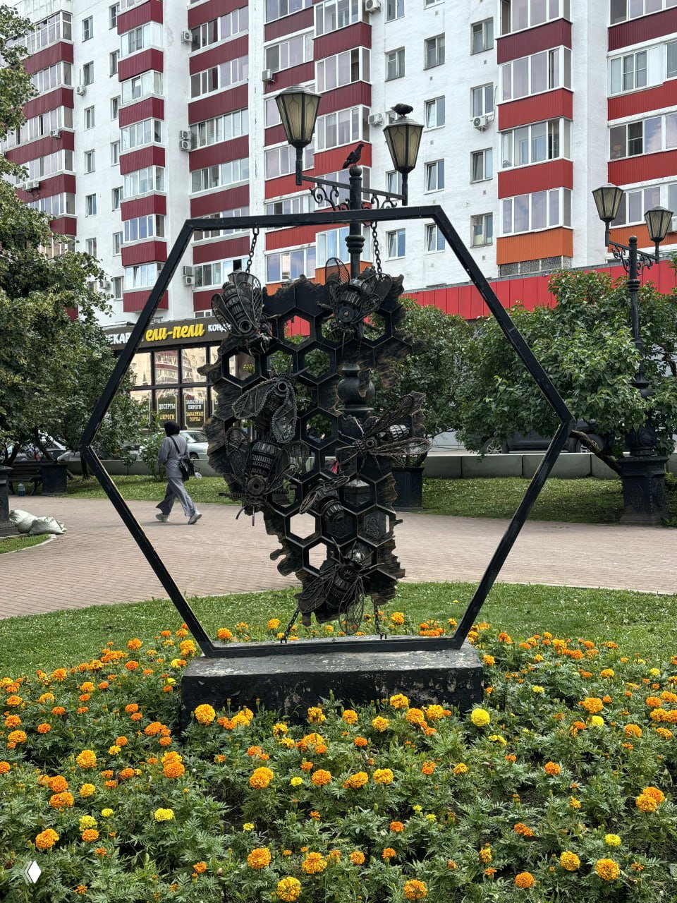 Городской сквер с художественной металлической инсталляцией в форме сот и клумбой с бархатцами на фоне жилого дома и фонарей.