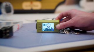 Рука держит портативную DIY-камеру ImageGenCam на столе: компактный корпус, небольшой LED-дисплей с превью, вокруг компоненты