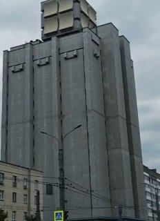 Высокое монолитное бетонное здание с крупной квадратной надстройкой на крыше, городской пейзаж с фонарём и жилыми домами на фоне пасмурного неба.