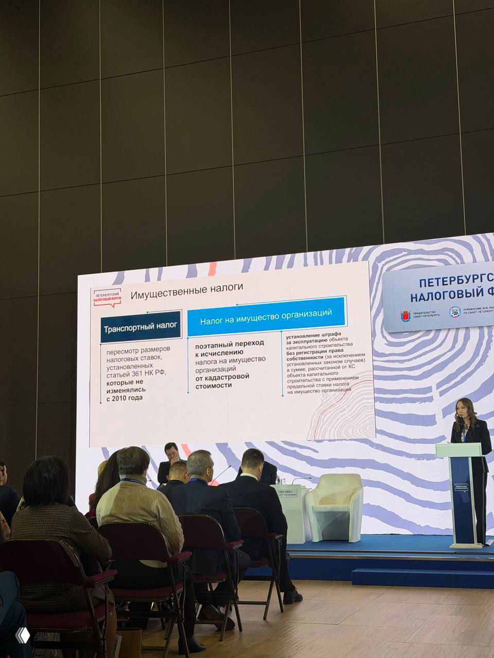 Фото сцены и экрана с темой «Имущественные налоги» на Петербургском налоговом форуме; спикер у трибуны и слушатели в зале.