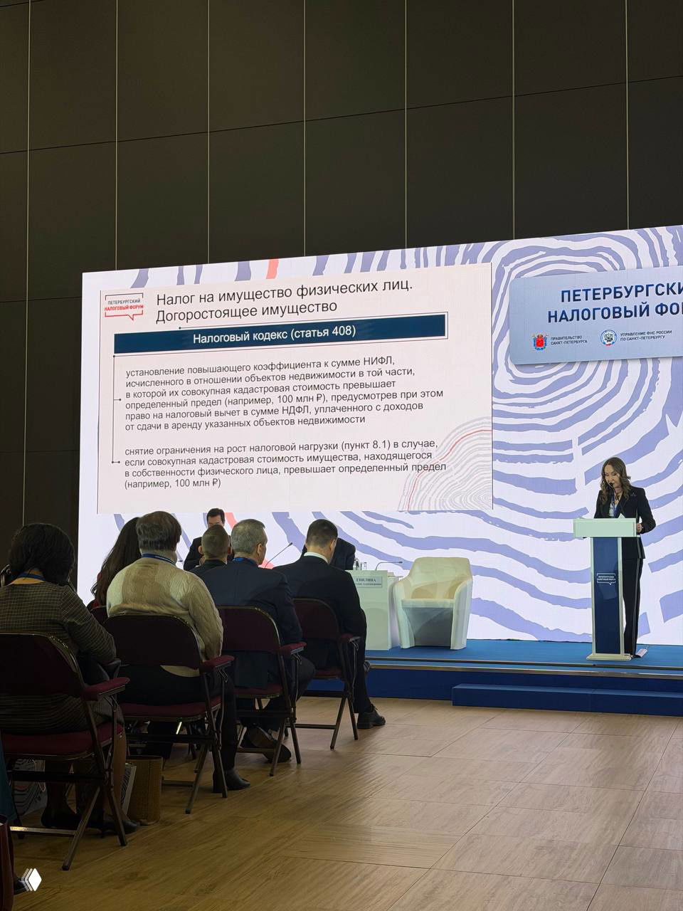 Кадр выступающей у трибуны на фоне слайда про налог на имущество физических лиц и сидящая аудитория в конференц-зале форума.