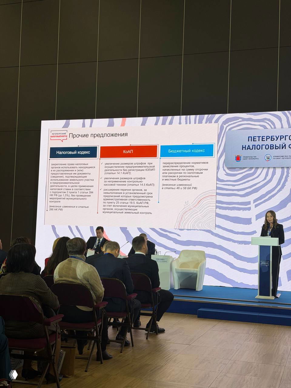 Изображение большого слайда с блоками предложений по Налоговому кодексу и аудитория конференции на Петербургском налоговом форуме.