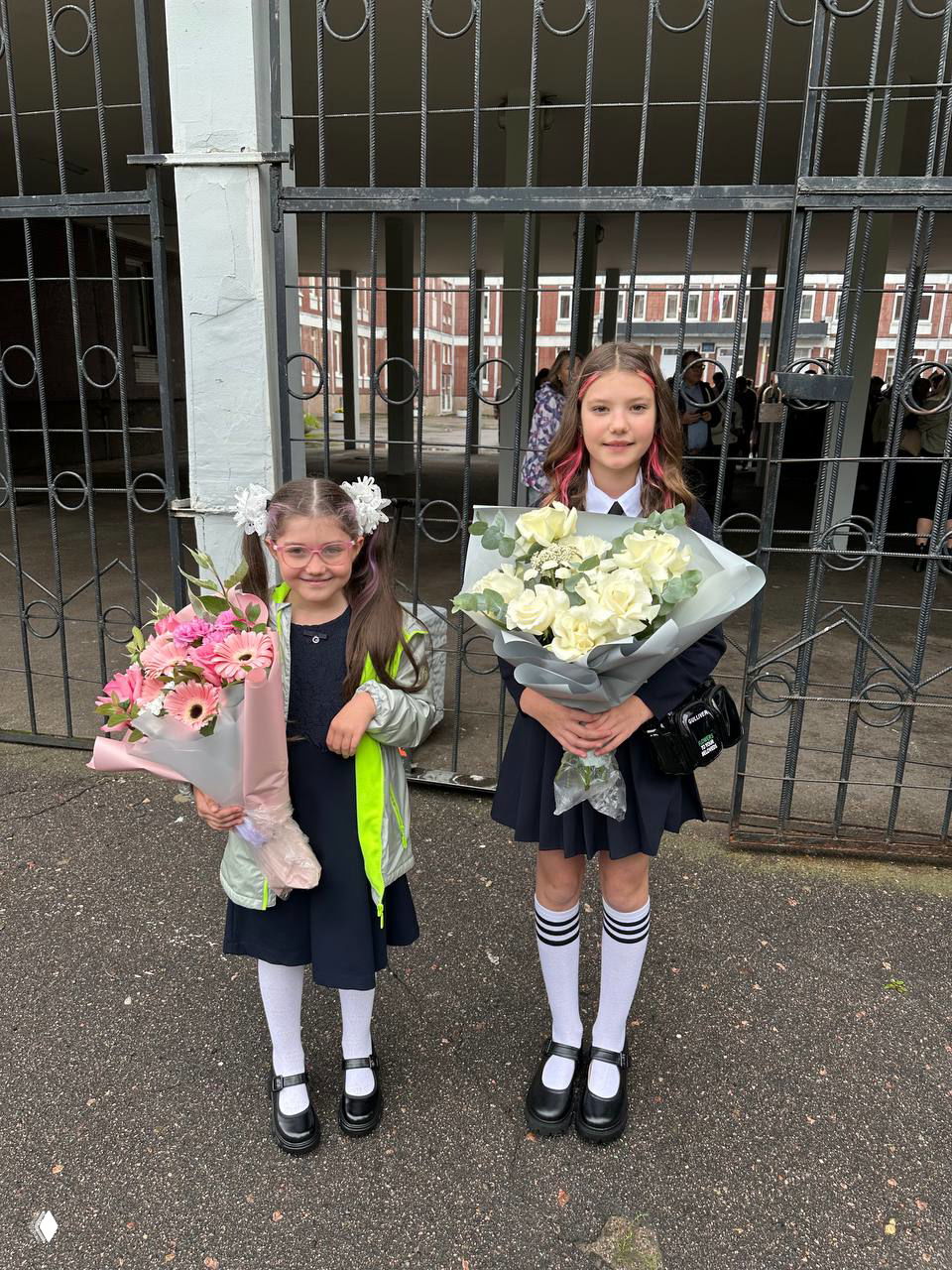 Две школьницы у ворот школы с большими букетами цветов: первая линейка, осенний школьный двор и радостный момент начала учёбы.