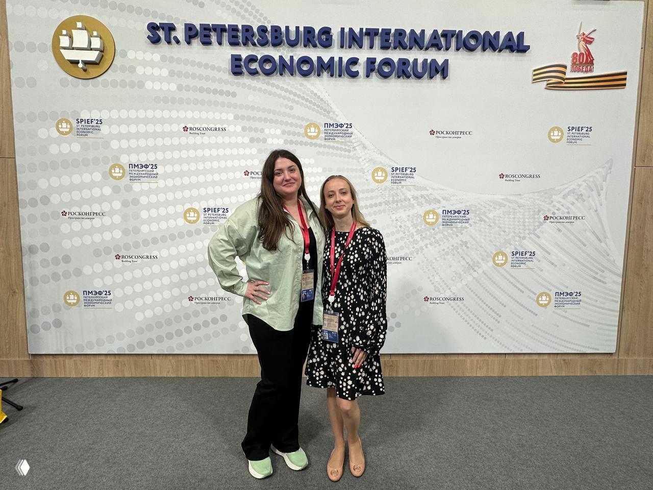 Две женщины на фоне баннера «ST.PETERSBURG INTERNATIONAL ECONOMIC FORUM» — фото с ПМЭФ или деловой площадки, бейджи на шее.